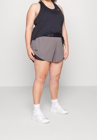 Persona con camiseta negra sin mangas, pantalones cortos deportivos grises, calcetines blancos y zapatillas blancas, de pie sobre un fondo simple.