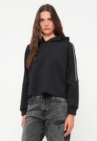 Svart kort hoodie med snøring i hetta, sidestriper og løs passform. Kombinert med slitte grå jeans, som gir et avslappet utseende.