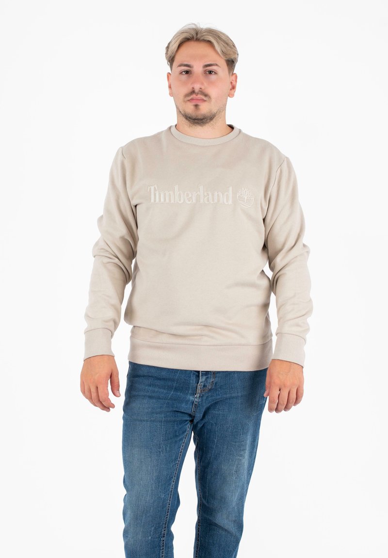 Felpa beige con logo "Timberland" ricamato, colletto rotondo, polsini e fondo a costine. Abbinata a jeans di denim blu.