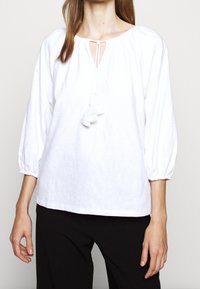Biała bluzka z rękawami 3/4 w kształcie puff, dekoltem w kształcie łezki oraz detalem w postaci frędzli. Tkanina ma subtelny wypukły wzór kwiatowy. Gładka faktura.
