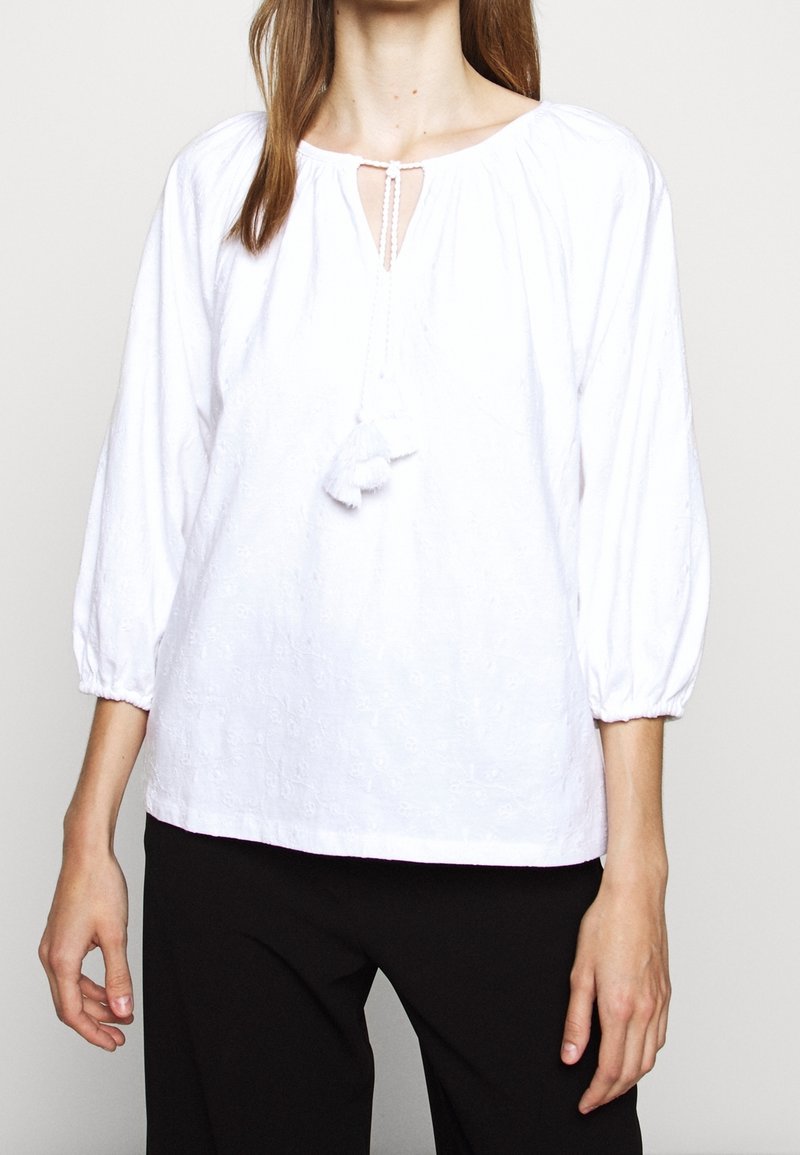 Biała bluzka z rękawami 3/4 w kształcie puff, dekoltem w kształcie łezki oraz detalem w postaci frędzli. Tkanina ma subtelny wypukły wzór kwiatowy. Gładka faktura.