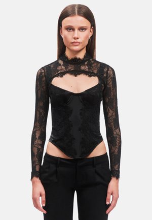 Donna con i capelli lisci che indossa un corsetto nero in pizzo a maniche lunghe e pantaloni neri, in piedi davanti a uno sfondo chiaro e uniforme.