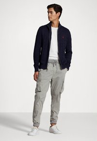 Μπλε σκούρα ζακέτα με πλέξη στην υφή πάνω από λευκό μπλουζάκι, συνδυασμένη με ανοιχτογκρι παντελόνι cargo και λευκά αθλητικά παπούτσια. Καθημερινό, εφαρμοστό σχέδιο με διακριτικές λεπτομέρειες.