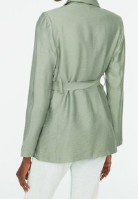 Vaaleanvihreä vyöllä varustettu takki, jossa on sileä pinta, istuva siluetti ja pitkät hihat. Sisältää halkion kauluksen ja takasauman yksityiskohtia.