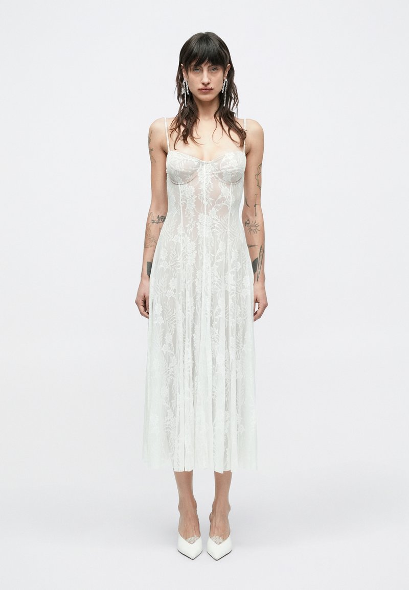 Biele kvetinové čipkované šaty s priliehavým korzetom a vzdušnou sukňou. Majú tenké ramienka a priehľadnú textúru, doplnené o špicaté biele lodičky.