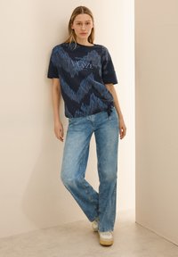 Camiseta azul marino con un patrón de zigzag en tonos más claros, combinada con jeans de corte recto de lavado medio y zapatillas blancas.