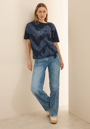 T-shirt bleu marine avec un motif en zigzag dans des tons plus clairs, associé à un jean droit de lavage moyen et des baskets blanches.