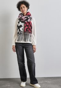 Strikkan scarf med et dristigt abstrakt mønster i rødt, pink og sort samt frynser. Modellen bærer en hvid top og mørke jeans, mens hun står op ad en lys væg.