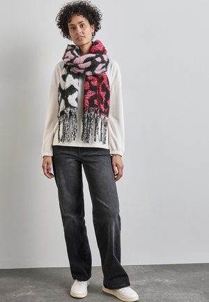 Strikkan scarf med et dristigt abstrakt mønster i rødt, pink og sort samt frynser. Modellen bærer en hvid top og mørke jeans, mens hun står op ad en lys væg.