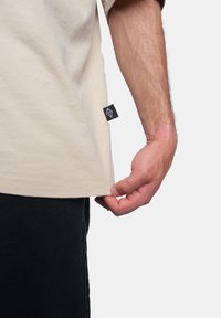 Béžové bavlněné tričko s krátkým rukávem, se zdobným prošíváním na lemu, bočním štítkem v černé barvě a přiléhavým střihem, zobrazené, jak je zataženo ve spodní části.