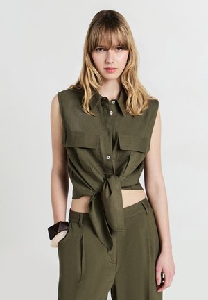 Giovane donna con capelli biondi che indossa una camicia senza maniche verde oliva annodata e pantaloni abbinati, in posa con una mano in tasca su uno sfondo neutro.