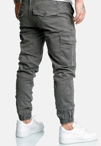 Mężczyzna w szarych spodniach cargo z elastycznymi mankietami i białych trampkach Nike Air, stojący na jednolitym tle.