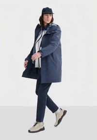 Voděodolný námořnicky modrý kabát s kapucí, se vzorovanou podšívkou, postranními kapsami a nastavitelnými manžetami, nošený přes lehký svetr a kalhoty.