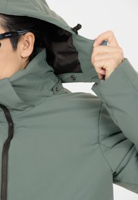 Giacca resistente all'acqua in un verde tenue, con colletto alto, zip frontale e cappuccio regolabile con fodera interna nera.