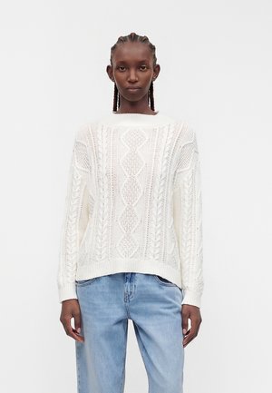 Kvinde med flettet hår iført en hvid kabelstrikket sweater og lyseblå jeans, stående mod en ensfarvet hvid baggrund.