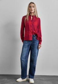 Camicia a quadri rossa con maniche lunghe e patta con bottoni, abbinata a jeans in denim blu larghi e sneakers bianche. Texture del tessuto liscia.
