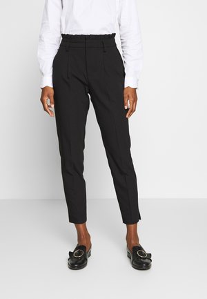 Person wearing black high-waist tapered pants with a paperbag waist, white long-sleeve shirt, and black slip-on shoes with gold buckle.