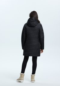 Schwarze, gepolsterte Jacke mit Kapuze, die ein vertikal gestepptes Muster aufweist. Getragen über schwarzen Leggings und hellbraunen Winterstiefeln.