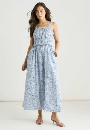 Woman standing wearing a sleeveless light blue floral maxi dress with smocked bodice and pink heeled sandals against a plain background.