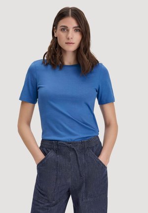 Frau mit schulterlangem braunem Haar, die ein blaues kurzärmeliges Shirt und dunkle Jeans-Hose mit Tunnelzug trägt und die Hände in den Taschen hat.