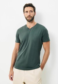 Tricou verde cu decolteu în V, fabricat dintr-un material moale, cu mâneci scurte, textura netedă și design ajustat, purtat cu pantaloni de culoare deschisă.