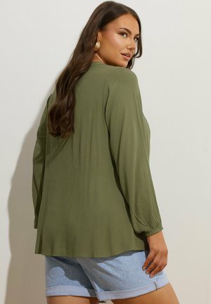 Frau mit langen braunen Haaren, trägt goldene Ohrringe, eine olivgrüne Langarmbluse und hellblaue hochgekrempelte Jeansshorts, von hinten gesehen.