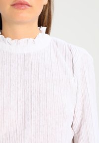 Blusa bianca con colletto alto a balze, caratterizzata da pieghe verticali e tessuto testurizzato. Maniche lunghe con una vestibilità leggermente ampia.