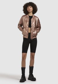 Broskvová bomber bunda s lesklým povrchem, žebrovaný límec a manžety, nošená přes černý crop top a cyklistické šortky, doplněná černými bojovými botami.