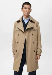 Beige trenchcoat met een dubbelzijdig ontwerp, donkere knopen, brede revers en manchettenaccenten. Draagt over een donker shirt en broek.