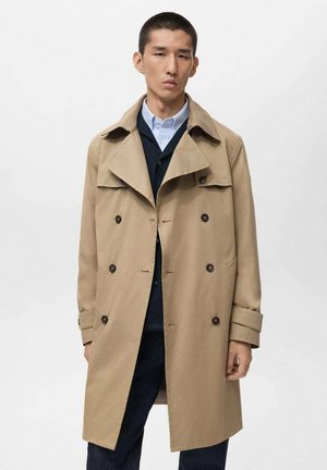 Trench de color beige con diseño de doble botonadura, botones oscuros, solapas anchas y acentos en los puños. Llevado sobre una camisa oscura y pantalones.