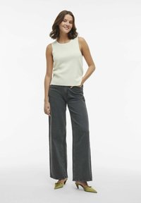 Top sin mangas de canalé en beige claro combinado con unos vaqueros oscuros grises de pierna ancha y cintura alta. La modelo lleva zapatos verdes con un pequeño tacón. Estilo sencillo.