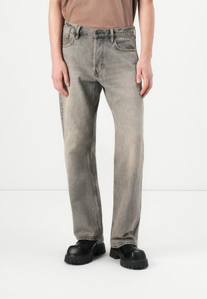 Person som har på seg falmede grå jeans med rett ben, svarte sko med tykk såle, og en brun topp foran en ensfarget hvit bakgrunn.