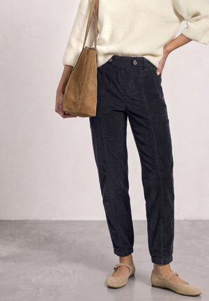 Personne portant un pantalon bleu foncé et un pull crème, avec un sac à bandoulière beige, et des chaussures Mary Jane beiges, debout sur un sol gris.