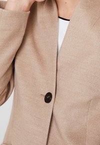 Beige uldblanding jakke med en enkelt sort knaplukning og klippet krave. Blød tekstur med minimal syning.