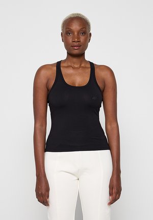 Débardeur noir ajusté sans manches et pantalon blanc portés par une personne debout aux cheveux blonds courts sur un fond uni.