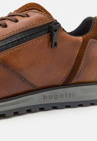 Sneaker in pelle marrone con superficie testurizzata, dettaglio della cerniera nera, suola grigia e "bugatti" impresso sul lato. Cuciture rinforzate visibili.