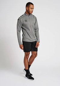 Veste de sport grise avec fermeture éclair sur le devant, col montant et bandes noires sur les épaules, assortie à un short noir et des chaussures de sport noires.