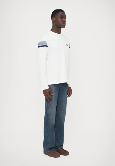 Άσπρο πουκάμισο με μακριά μανίκια και μπλε ρίγες στα μανίκια, συνδυασμένο με ανοιχτό μπλε τζιν και καφέ παπούτσια με κορδόνια, στέκεται σε απλό φόντο.