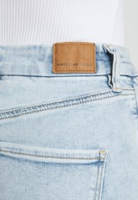 Ljusblå denimjeans med en läderlapp märkt "AMERICAN EAGLE" vid midjan. Har standardfickor och sydda sömmar.