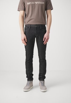 Homme portant un jean noir ajusté, un t-shirt taupe Emporio Armani et des baskets gris clair, debout devant un fond blanc uni.
