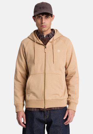 Jonge man met een beige zip-up hoodie, een donker geruit overhemd daaronder, een donkere spijkerbroek en een grijze pet, staand tegen een effen lichte achtergrond.