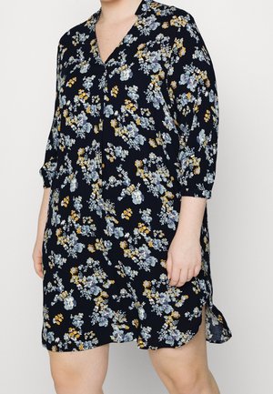 Person iført en mørkeblå blomstret kjole med gule og hvide blomster, trekvartærmer og en sideslidse, stående mod en neutral baggrund.