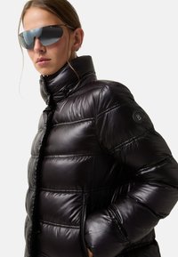 Schwarze Pufferjacke mit hohem Kragen, glänzender Textur und horizontalem Steppmuster; verfügt über ein Logo-Patch am Ärmel und Reißverschlusstaschen.