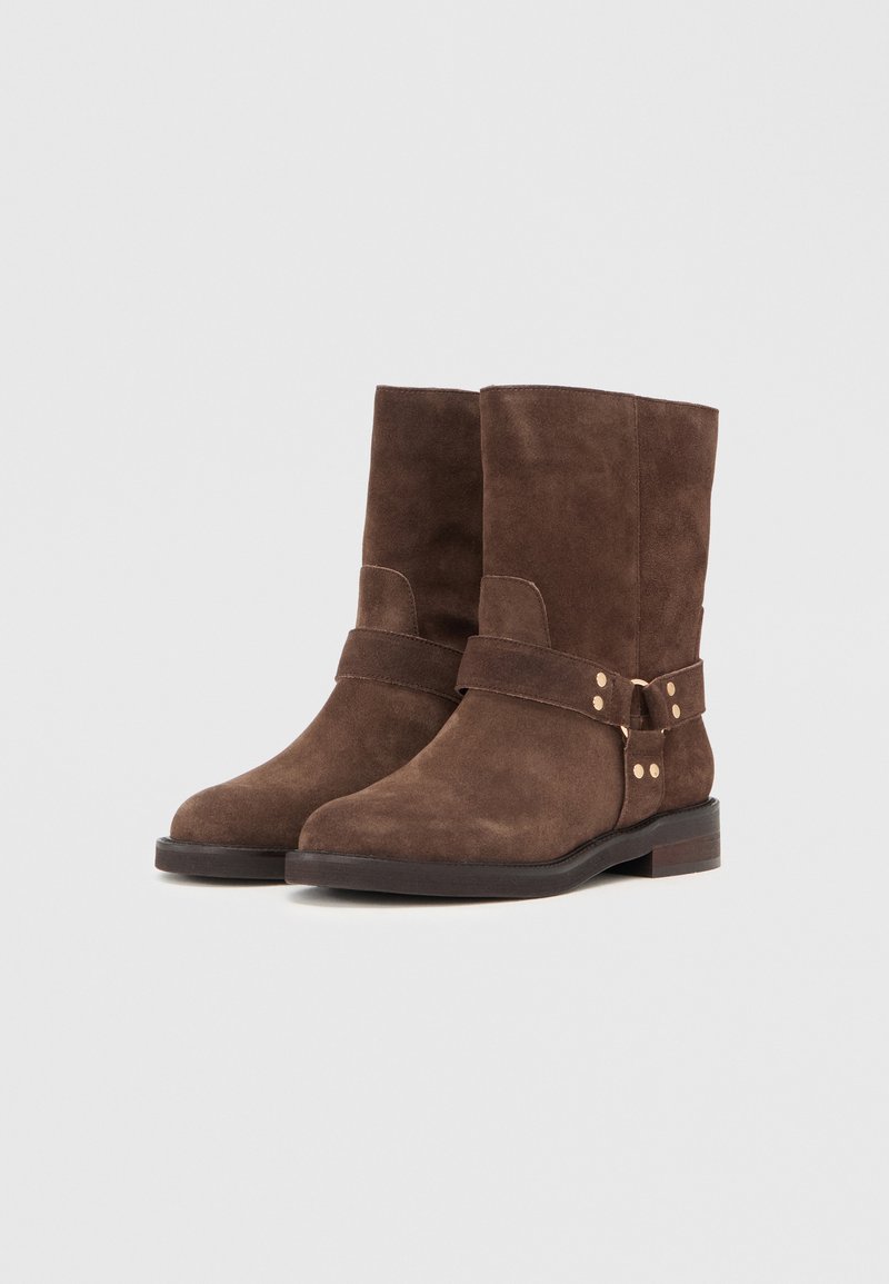Braune Wildleder-Stiefeletten mit flacher Sohle, ausgestattet mit einem breiten Riemen und goldfarbenen Nietenakzenten rund um den Knöchel.