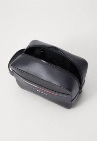 Čierna syntetická toaletná taška s zakriveným dizajnom, zipsom a jedným uchom. Obsahuje horizontálne pruhy a logo na prednej strane.