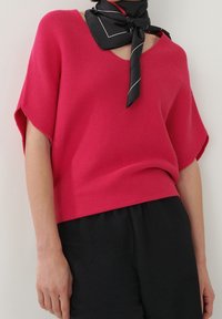 Růžový pletený top s výstřihem do V, širokými rukávy a volným střihem. V kombinaci s černými kalhotami a černým šálou s bílými doplňky.