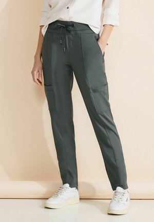 Frau trägt dunkelgrüne Cargo-Hose mit Kordelzugbund und weißen Turnschuhen vor beigefarbenem Hintergrund.