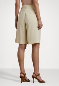 Bežid kõrge vöökohaga lühikesed püksid, millel on lahtine istuvus, sile tekstuur, lame eesmine osa ja kaks tagumist taskut. Selga pandud pruuni rihmaga kontsakingadega.