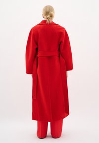 Cappotto rosso in lana con vita a cintura, maniche ampie e colletto, caratterizzato da una silhouette lunga e una texture liscia, indossato sopra pantaloni coordinati.