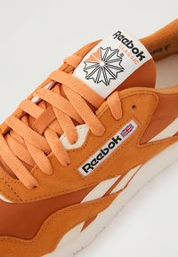 Reebok klassiske sneakers i oransje semsket skinn, med hvite detaljer, flate snørebånd og en logomerke med grafisk design på tungen.